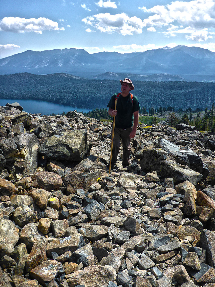 Hiker with brimmed hat and walking stick standing on talus with mountains in the distance