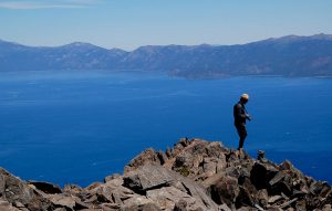 Man with camera and brimmed hat on mountain summit overlooking Lake Tahoe